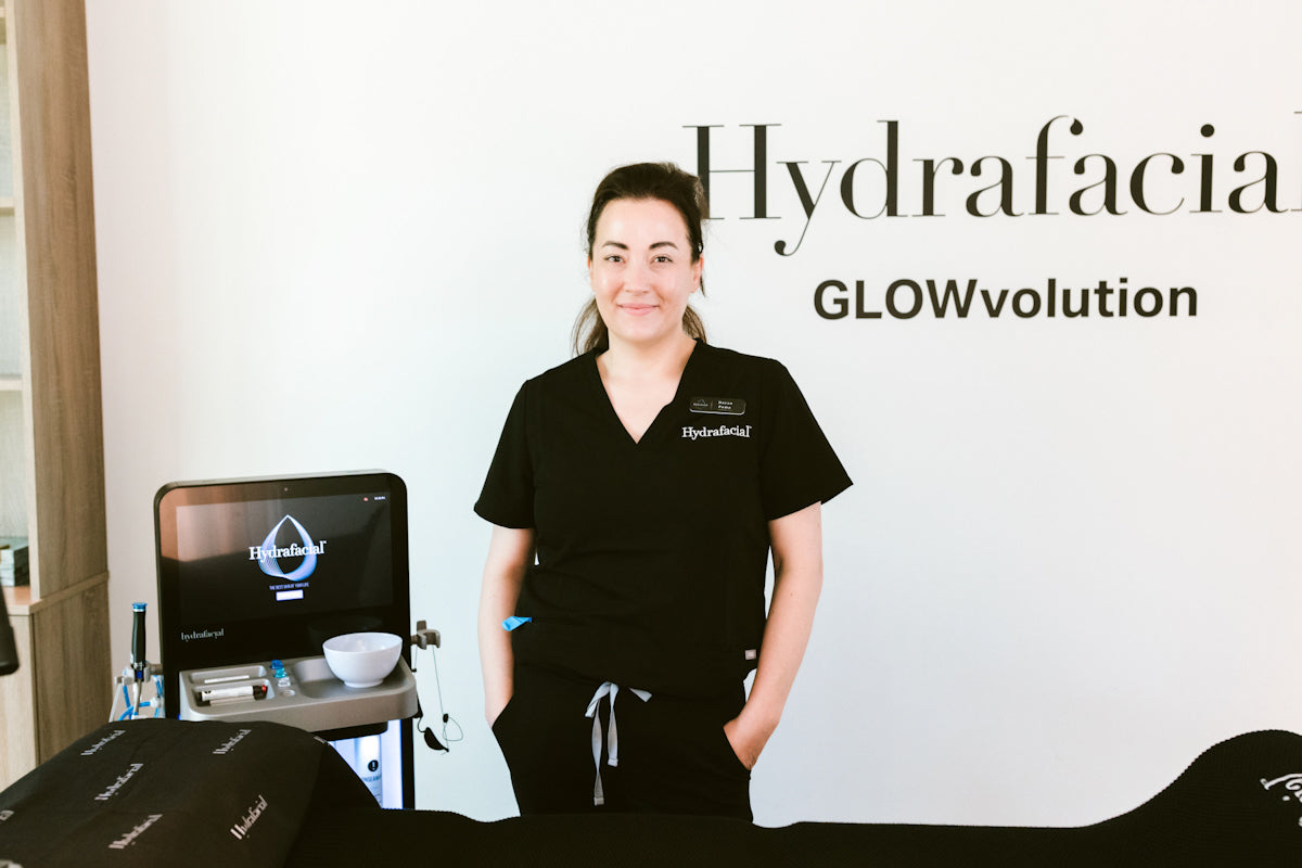 Doran Poma in black scrubs standing next to a Hydrafacial machine with 'Hydrafacial GLOWvolution' branding.