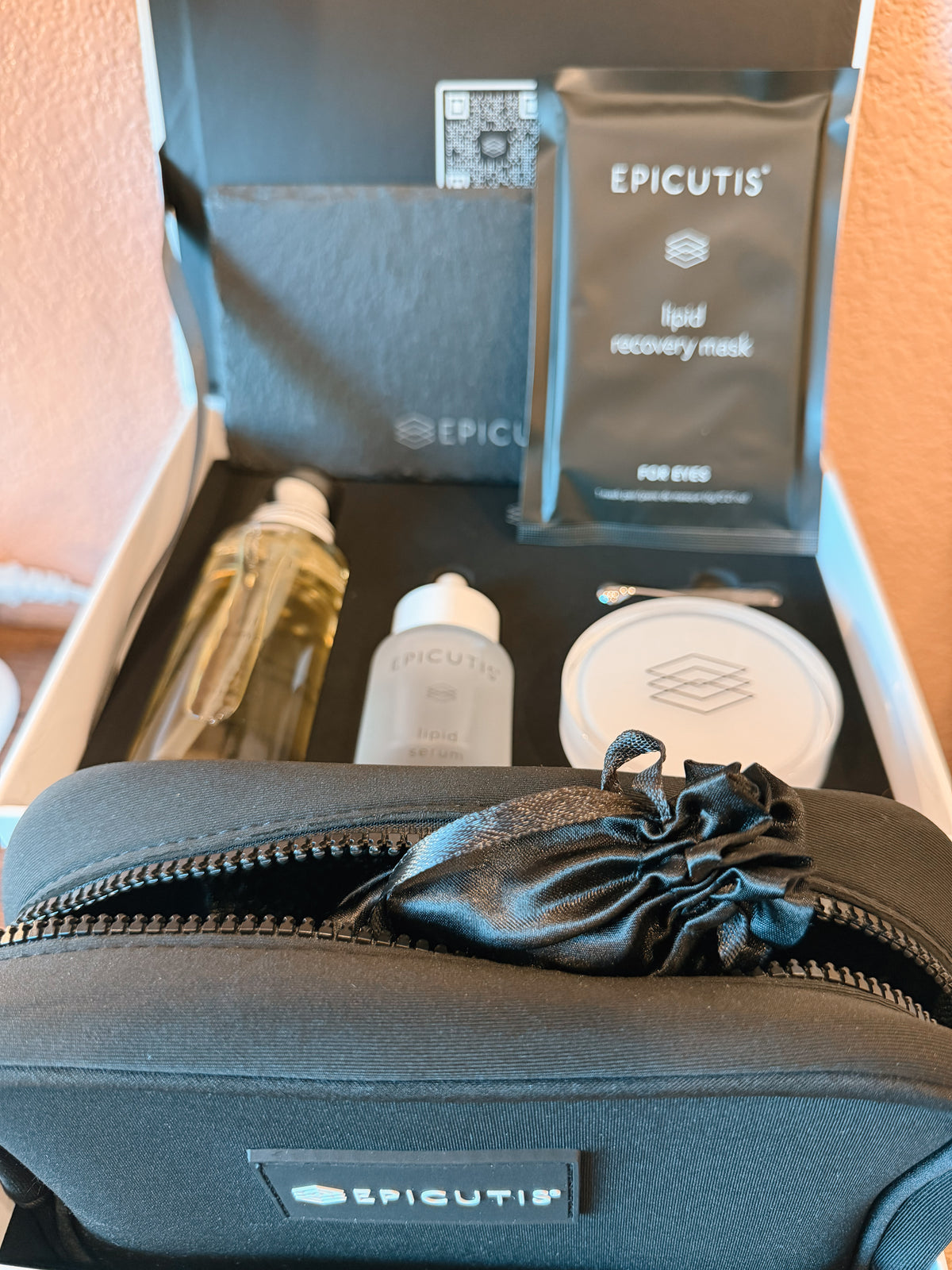 Epicutis skincare gift set displayed inside its open box, featuring a black zippered pouch with a satin drawstring, a black liquid recovery mask packet, a clear bottle with white cap, and other skincare items.