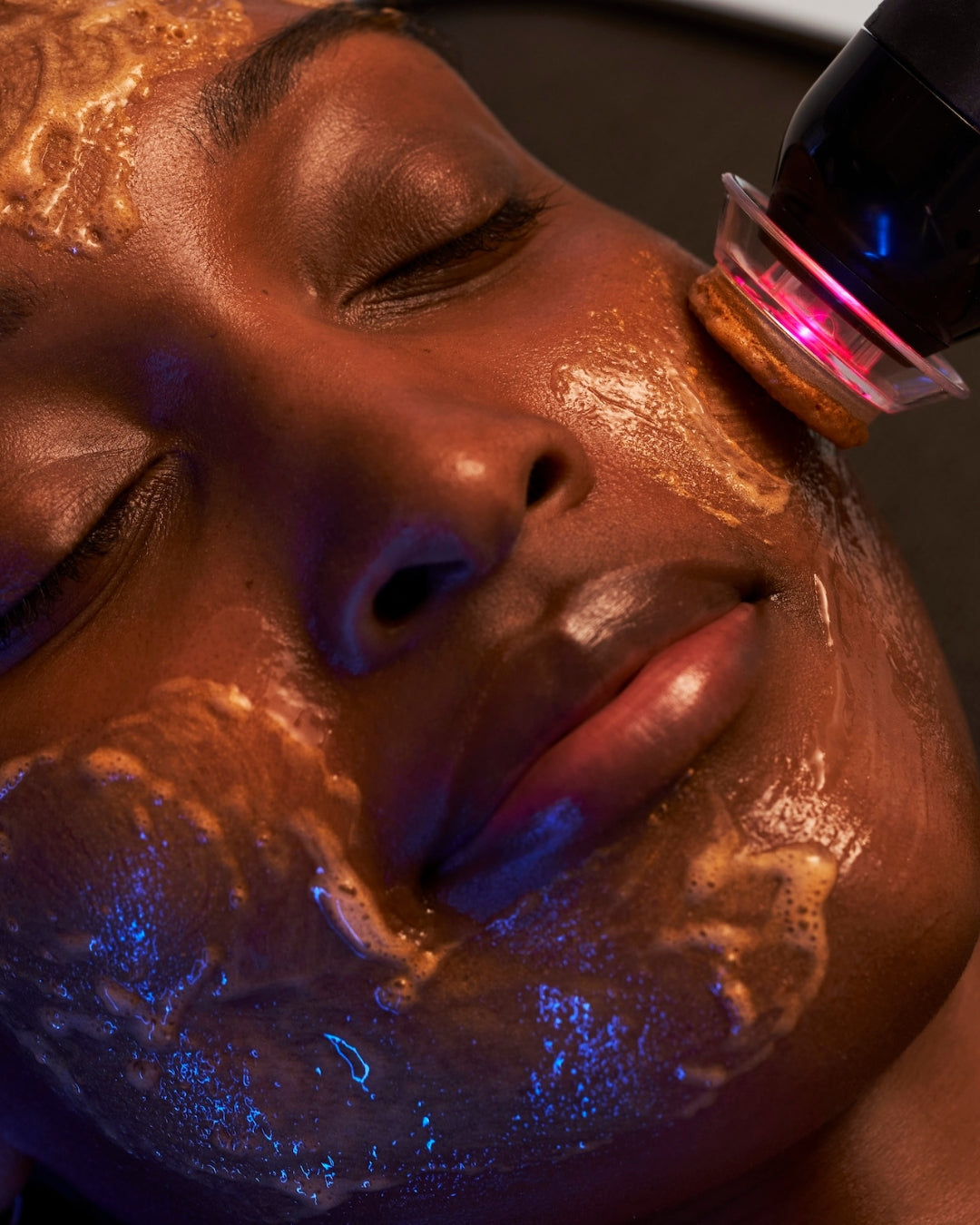 Close-up of a person's face with a glowing facial mask and gel applied, while a handheld device with a pink light emits blue and red illumination onto the skin. woman receiving a glo2facial.
