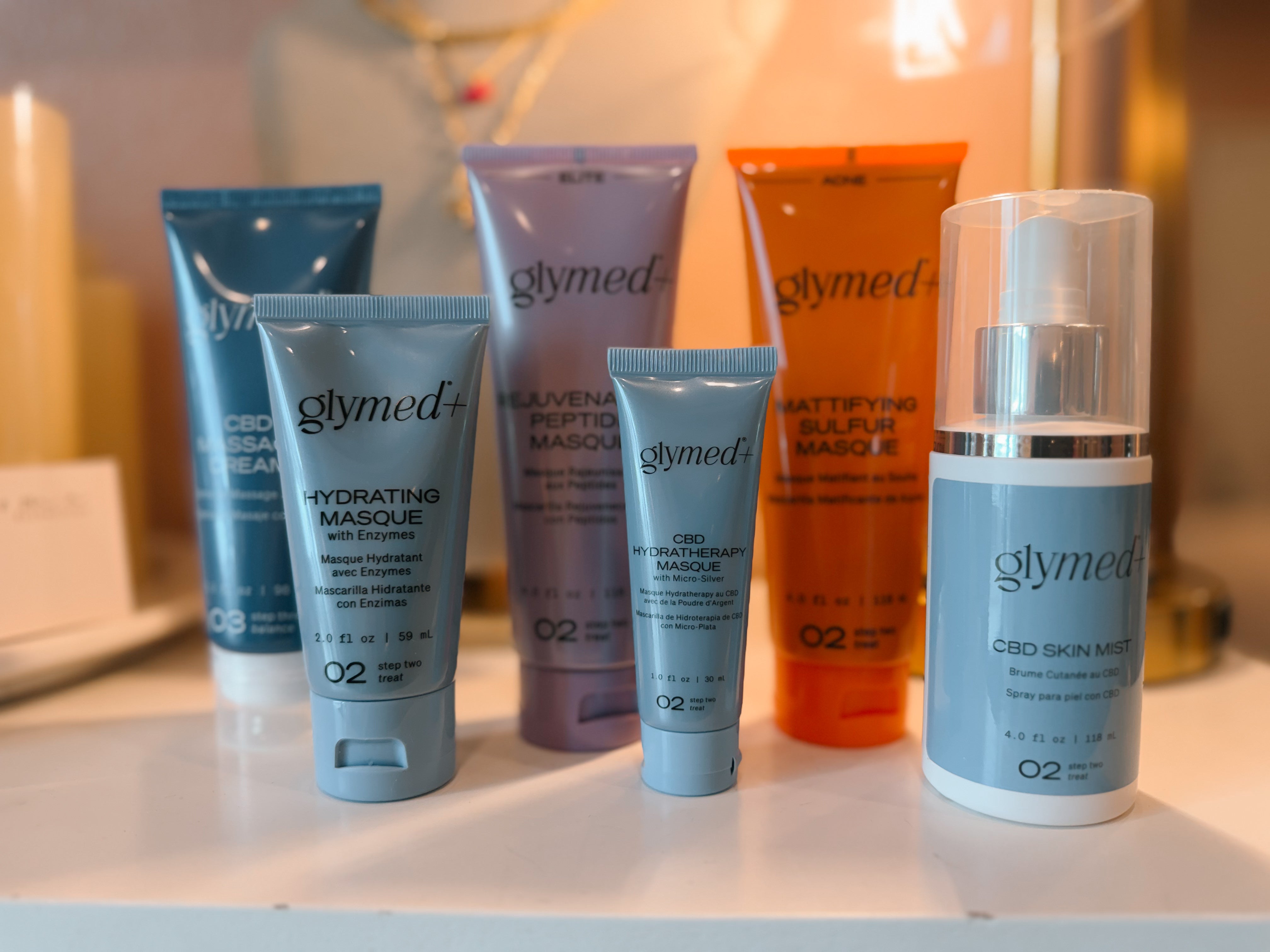 Glymed skincare products on a surface with a blurred background. Products from left to right CBD massage cream, hydrating masque, rejuvenating peptide masque, CBD hydratherapy masque, mattifying sulfur masque, glyemed CBD skin mist.