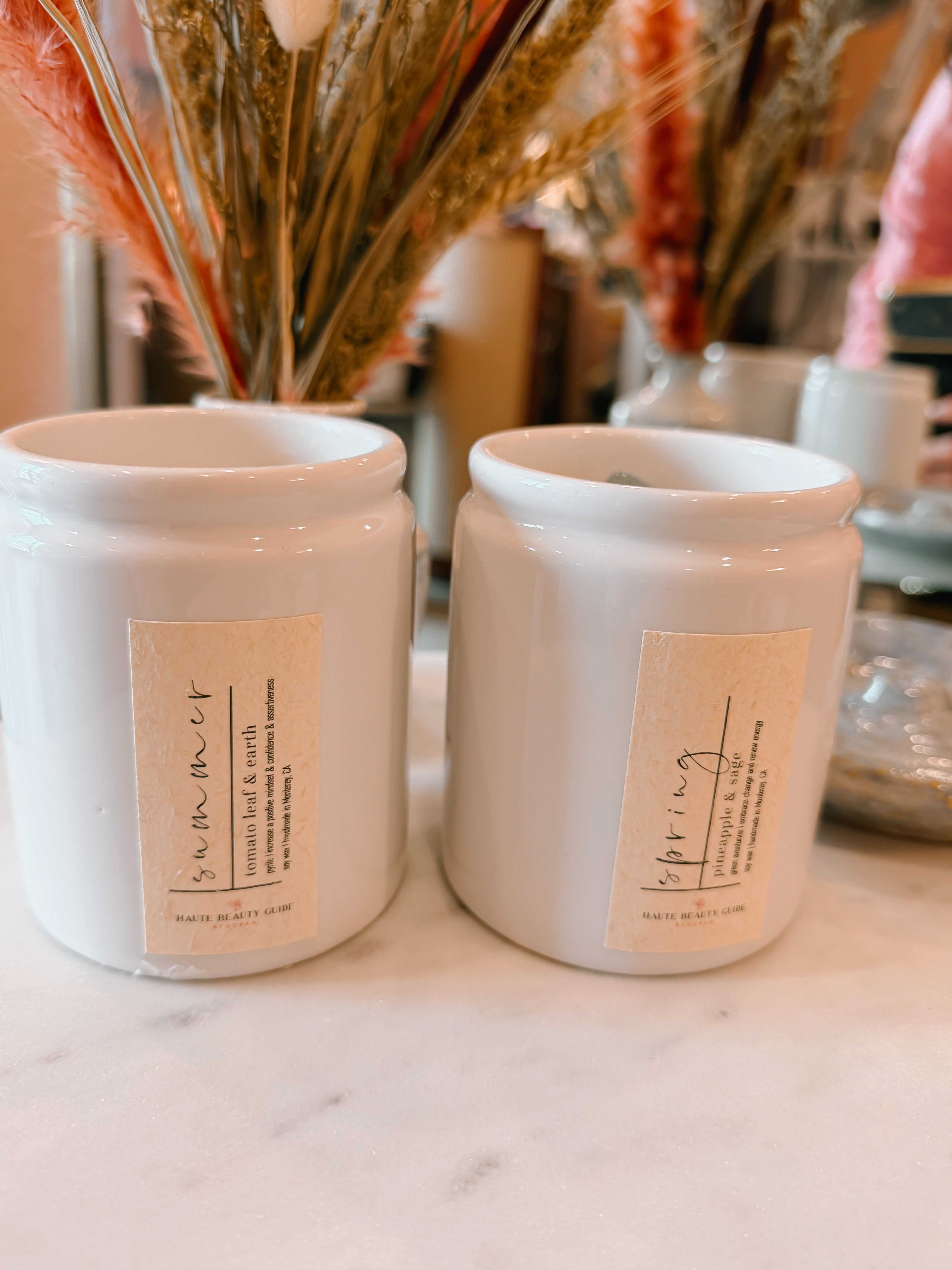 Two white ceramic containers with labels on a marble surface, with decorative plants in the background.