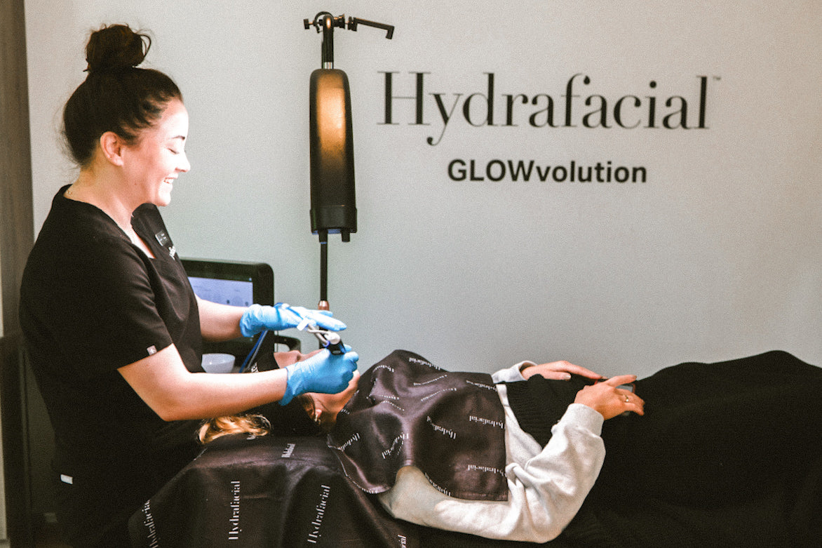 Doran Poma performing a Hydrafacial treatment on a woman laying in a bed. Hydrafacial logo and GLOWvolution logo in background.