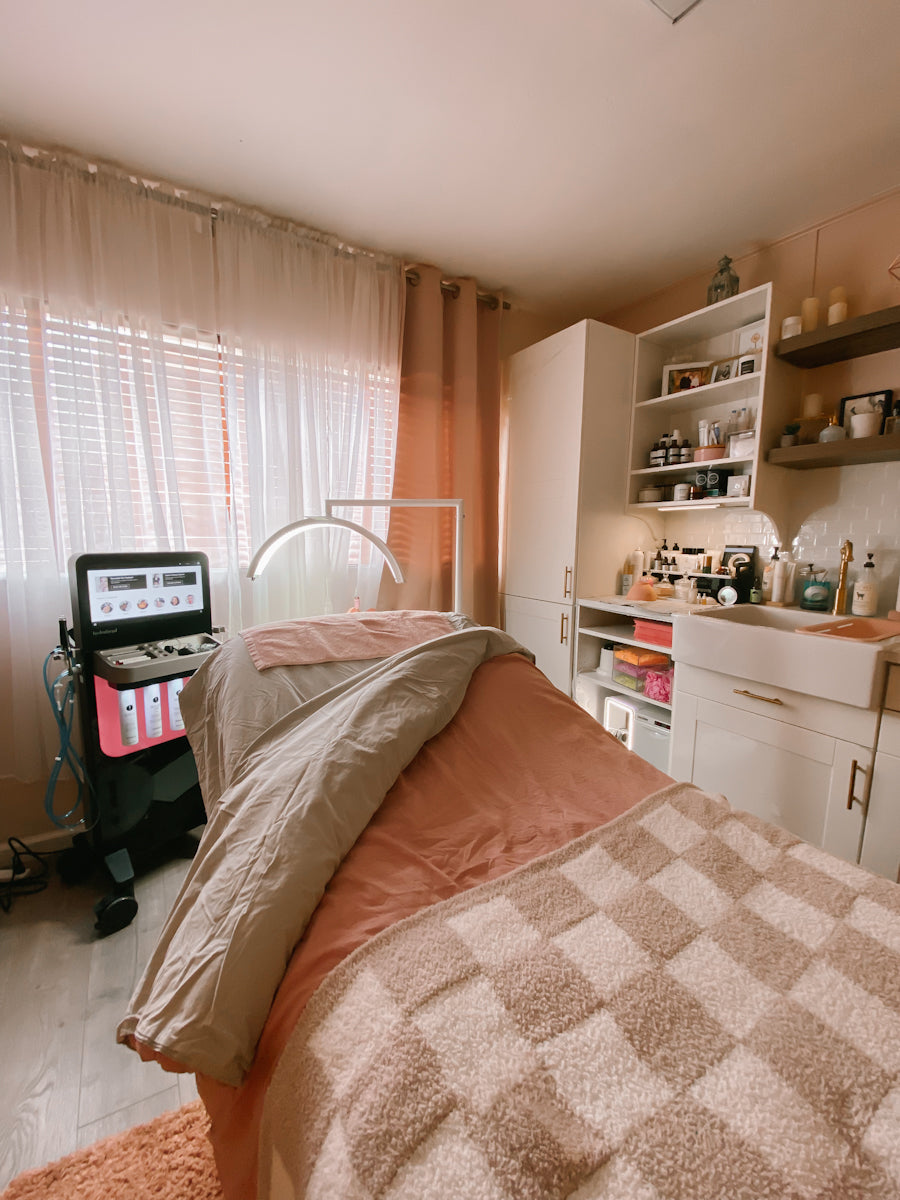 A spa treatment room featuring a treatment bed with peach and beige checkered blankets, a massage table machine, white cabinets, and shelves with skincare products.