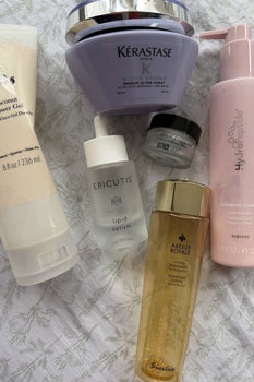 Flatlay of six empty beauty products on a floral fabric background: HydroPeptide Cashmere Cleanse Facial Rose Milk, Kérastase Blond Absolu Masque Ultra-Violet hair mask, PCA Skin Ideal Complex Restorative Eye Cream, Guerlain Abeille Royale Fortifying Loti