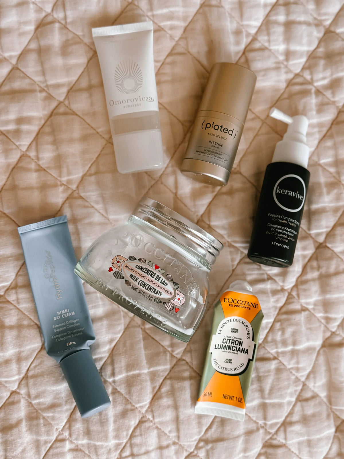 a bundle of empty skincare products on a pink blanket.  products include Plated skin, Hydropeptide Nimni Day, L'occitain body and hands creams, Omorovicza face wash and Hydrafacial Keravive leave on spray