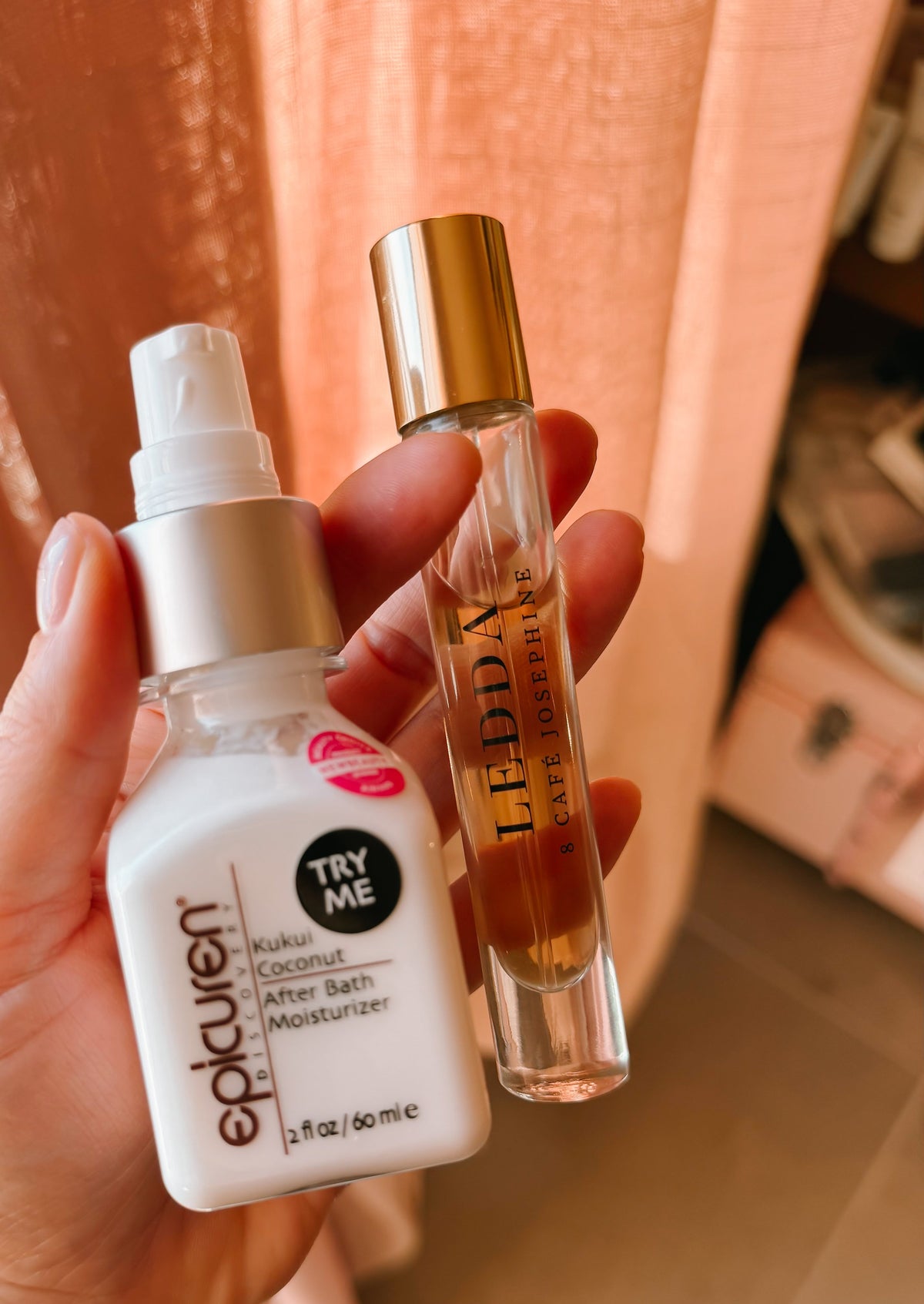 Hand holding a LEDDA 8 Café Josephine rollerball perfume and Epicuren's Kukui Coconut After Bath Moisturizer against a pink curtain backdrop.
