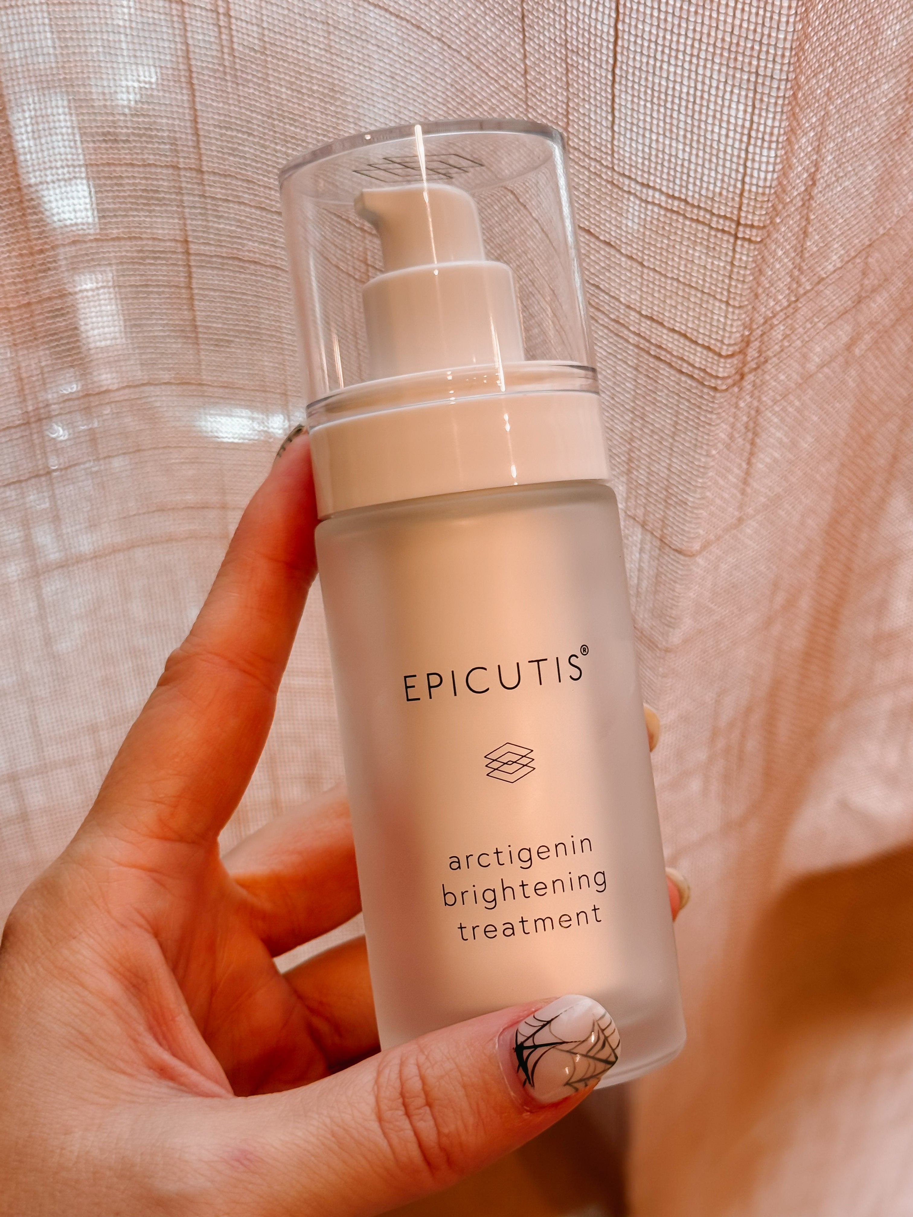 hand holding the epicutis arctigenin brightening treatment in Marina, California.  womans hands have halloween cobweb painted nails.  A pink mesh curtain is behind the bottle. 