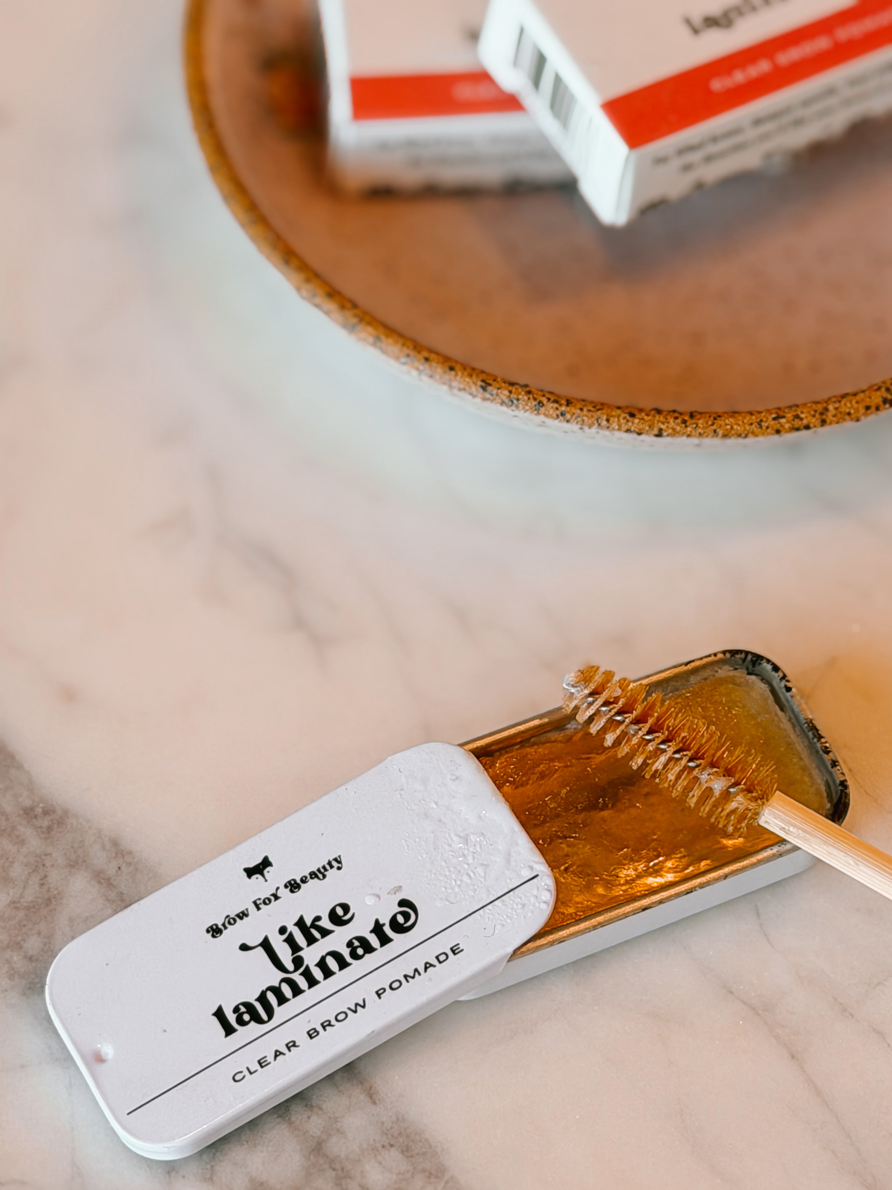 Clear brow pomade in a metal tin with a brush on a marble surface