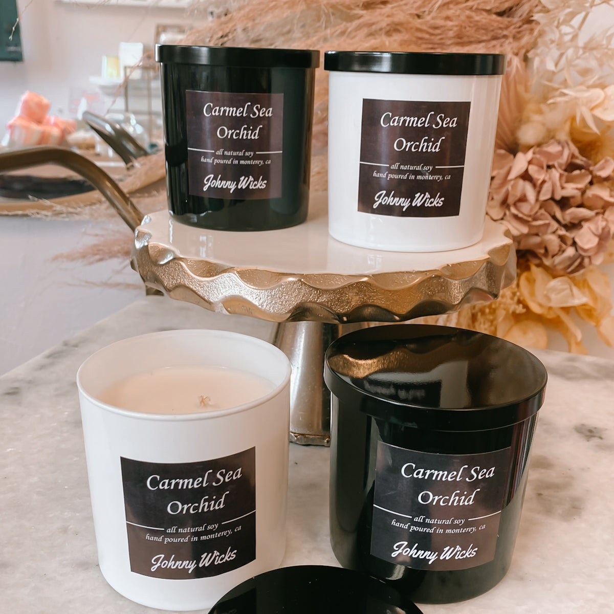 Four 'Carmel Sea Orchid' candles by Johnny Wicks on a decorative stand with flowers in the background.