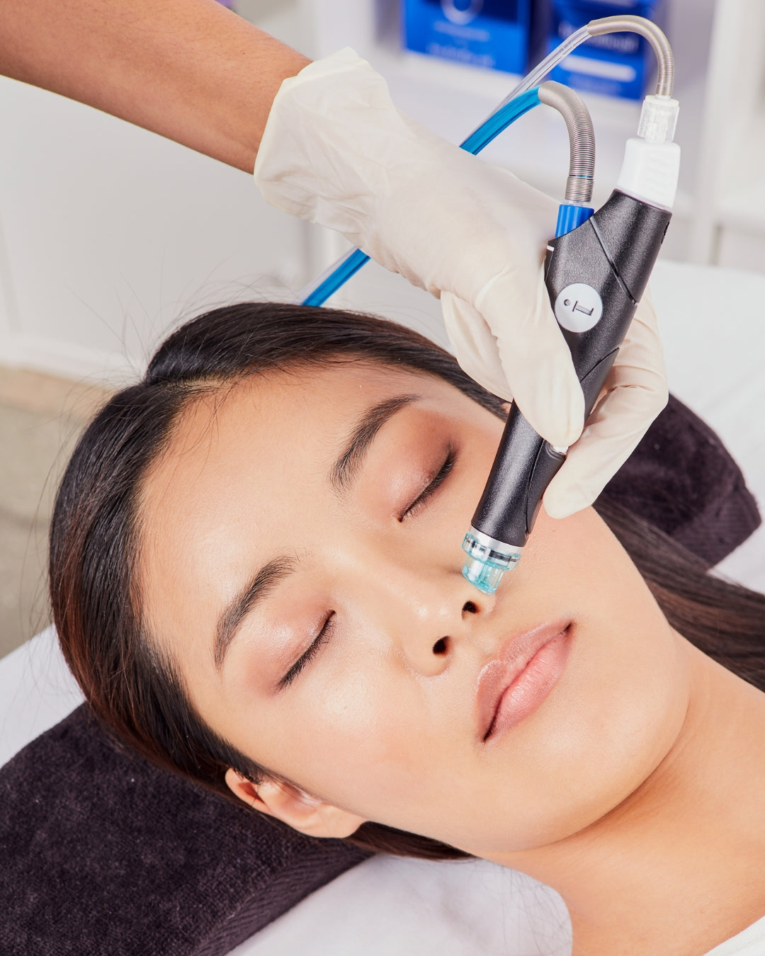 A close-up of a skincare treatment in progress, showing a person lying down with their eyes closed while a gloved hand holds a handheld device against their nose area. The device is black and silver with blue accents.
