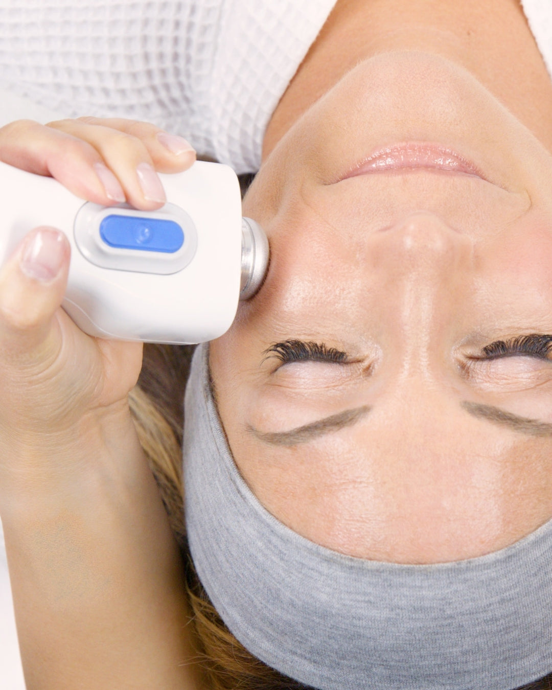 A close-up of a woman lying down with her eyes closed, using a white handheld facial device with a blue button on her forehead and temple. She is wearing a gray headband. She is receiving the glacial glide,