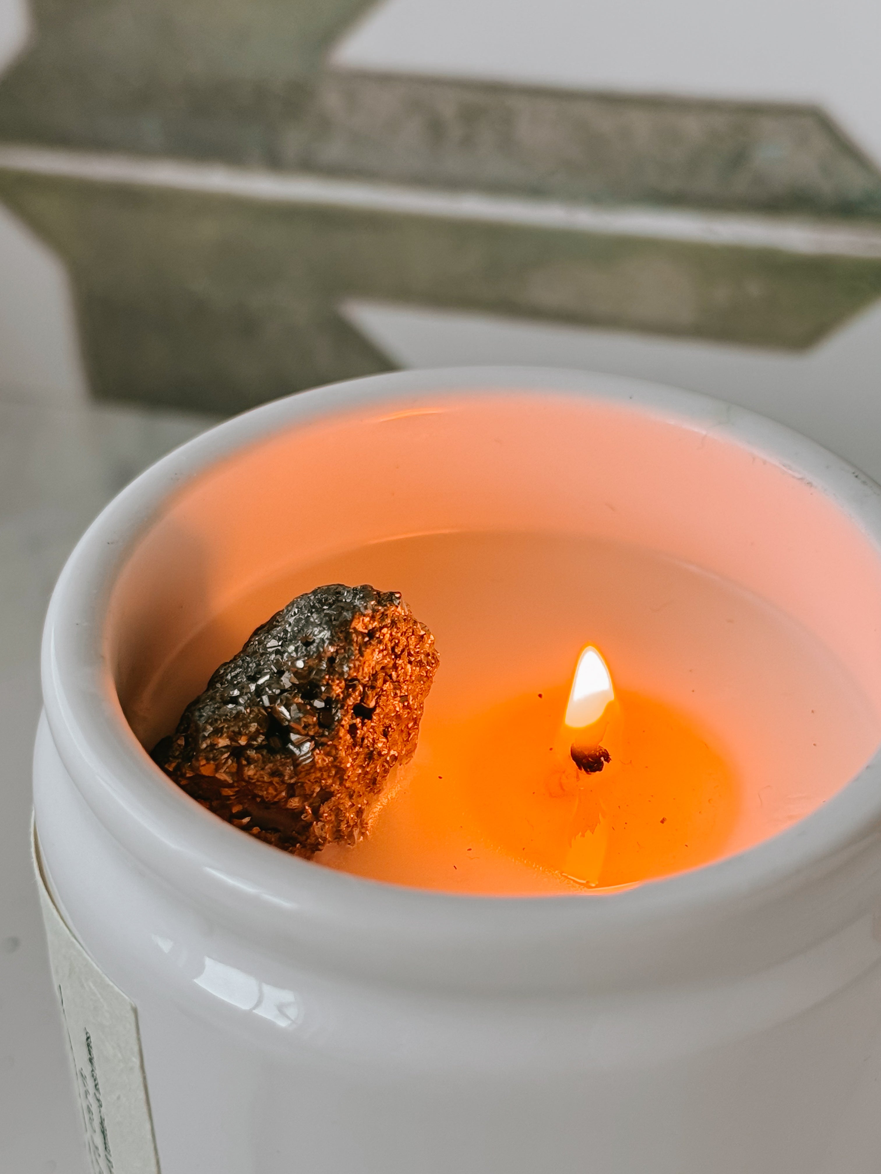 Candle in a white container with a small flame and dark pyrite rock inside, on a light background.