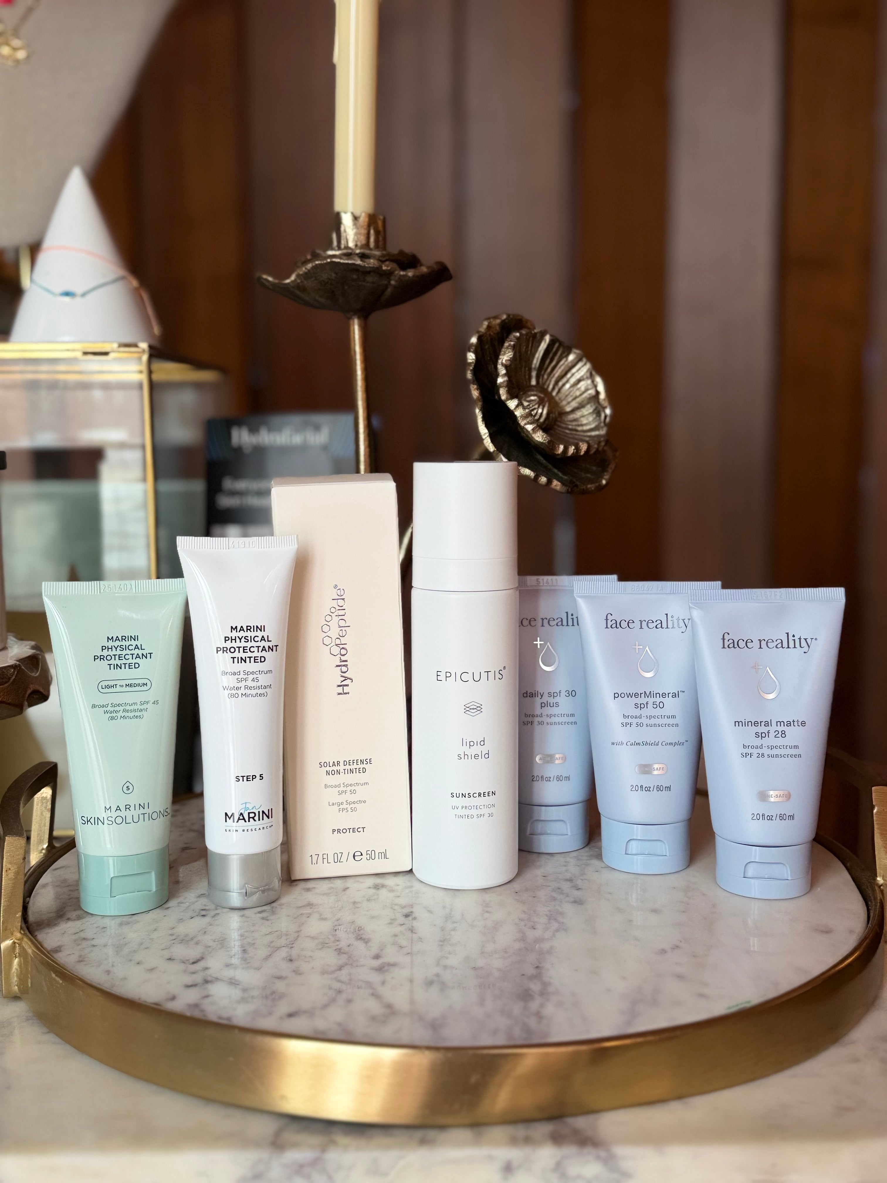 Collection of skincare products on a marble tray with decorative items in the background.  Products from left to right Marini Physical Protectant Tint Light to Medium, Marini Physical Protectant Tinted SPF 45, Hydropeptide Solar Defence non-tinted SPF, Epictuis Lipid Shield SPF 30, Face Reality Daily SPF 30 Plus, Face REality PowerMineral SPF 50, Face Reality mineral matte spf 28